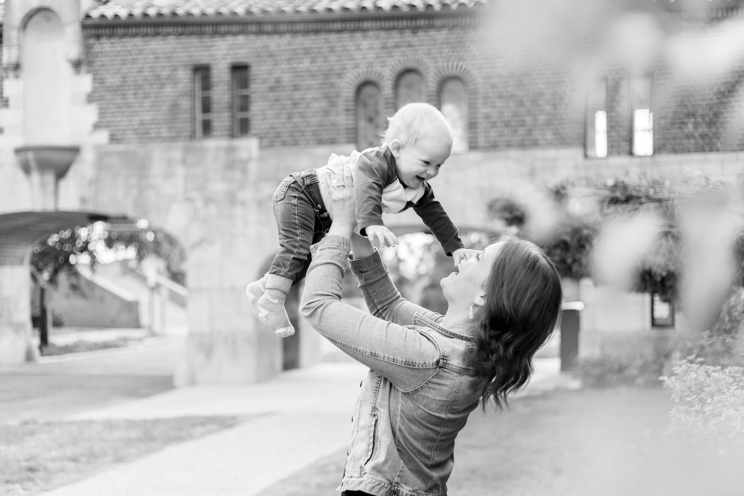 Parent lifts baby in air while playing outdoors near historic building.