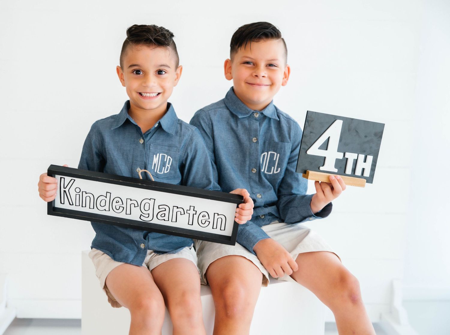 Two young twin brothers in matching chambray shirts holding Kindergarten and 4th grade signs.