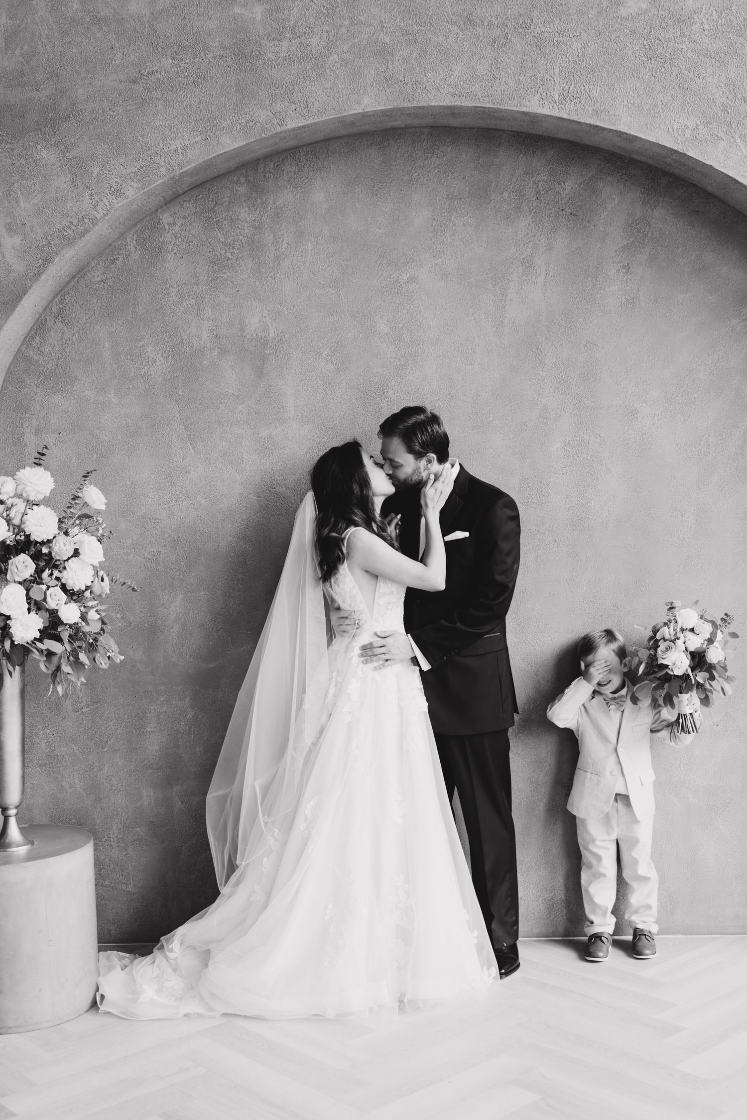 A sweet wedding moment captured in black and white, with a young ring bearer peeking from behind floral arrangements.