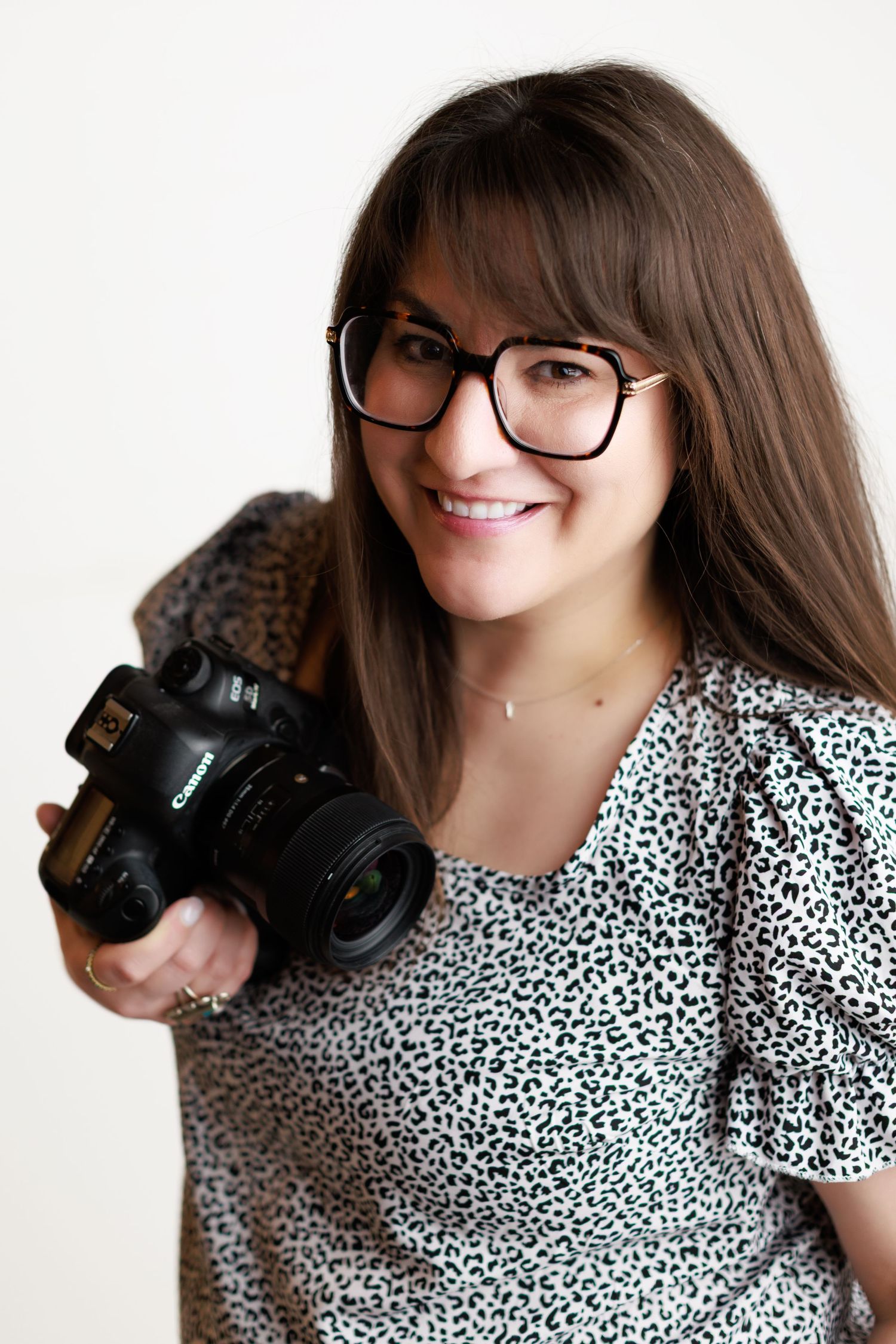 A photographer wearing glasses and a patterned blouse holds a professional DSLR camera against a white background.