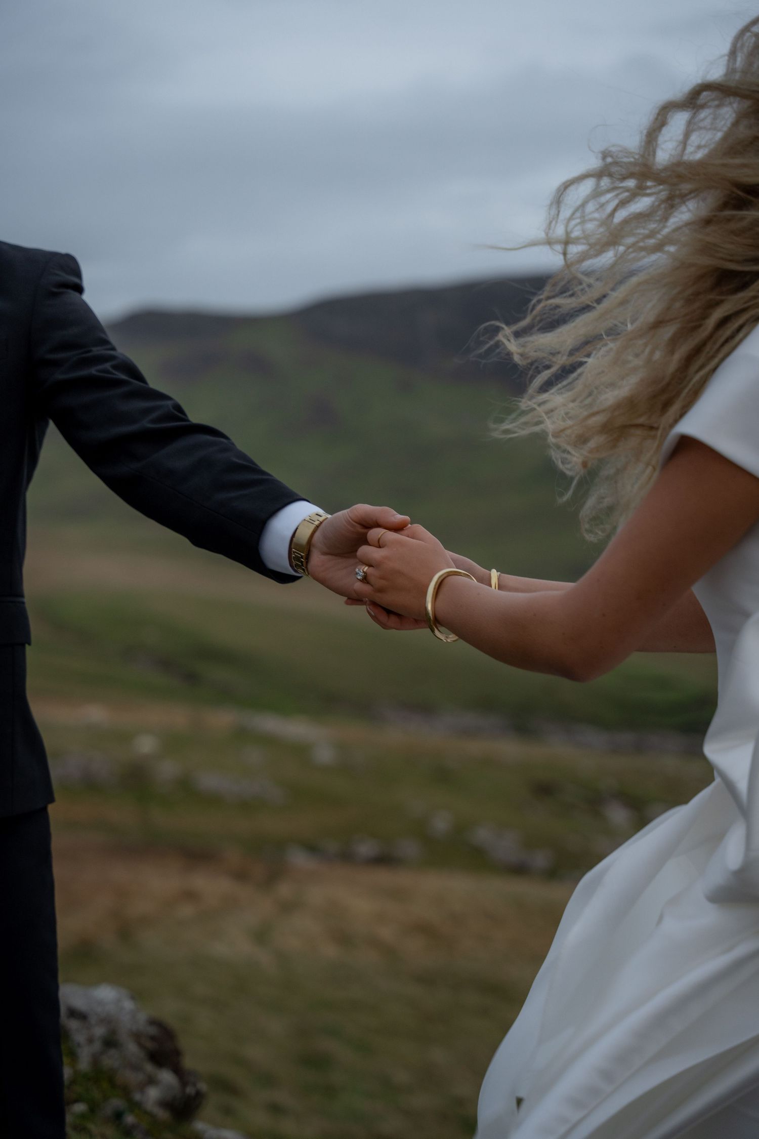 A newlywed couple holds hands on the Isle of Skye, surrounded by rugged cliffs and misty skies. Captured by Kaci, NYC wedding photographer.