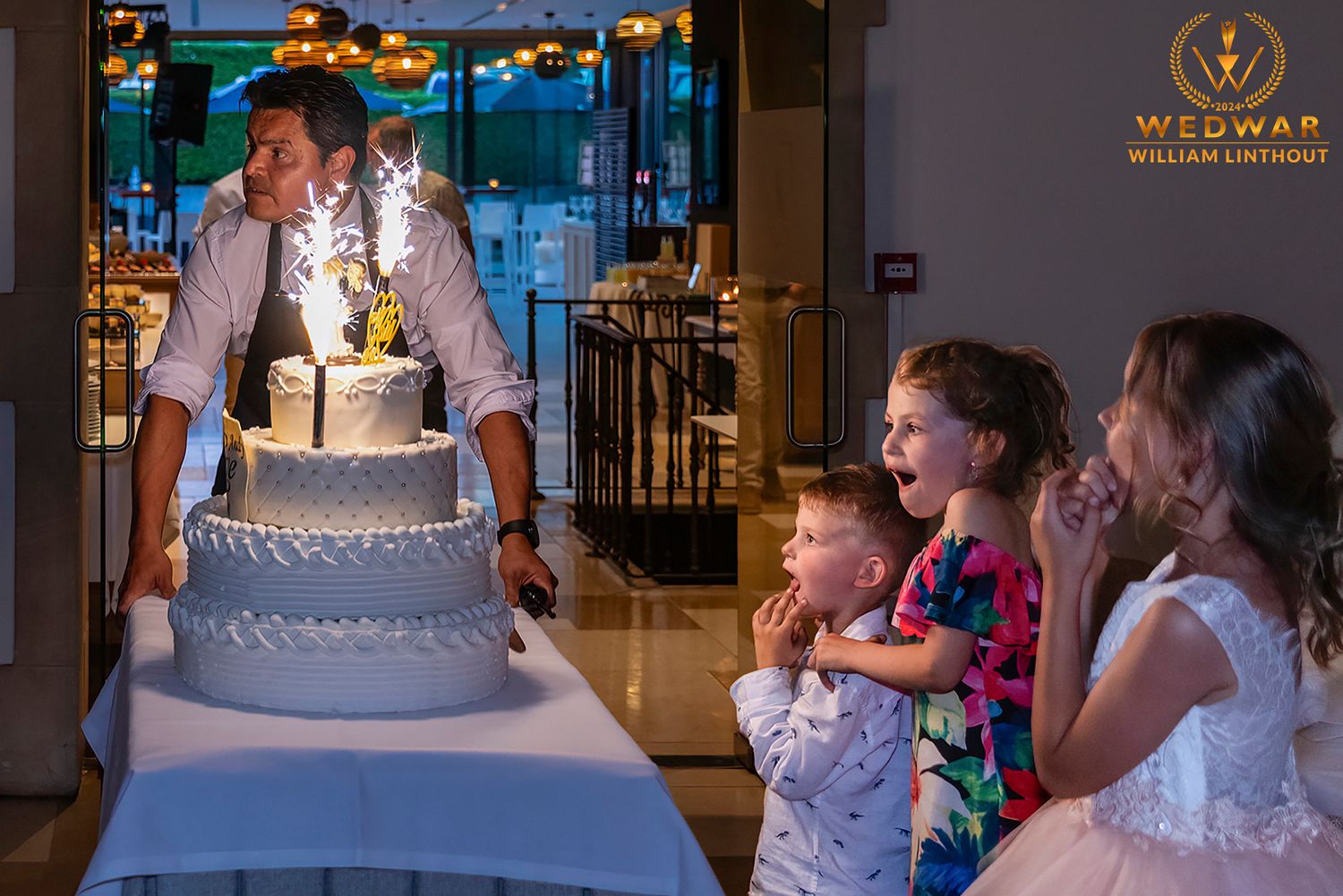 Children watch in delight as sparklers illuminate an elegant tiered wedding cake in a beautifully lit reception venue.