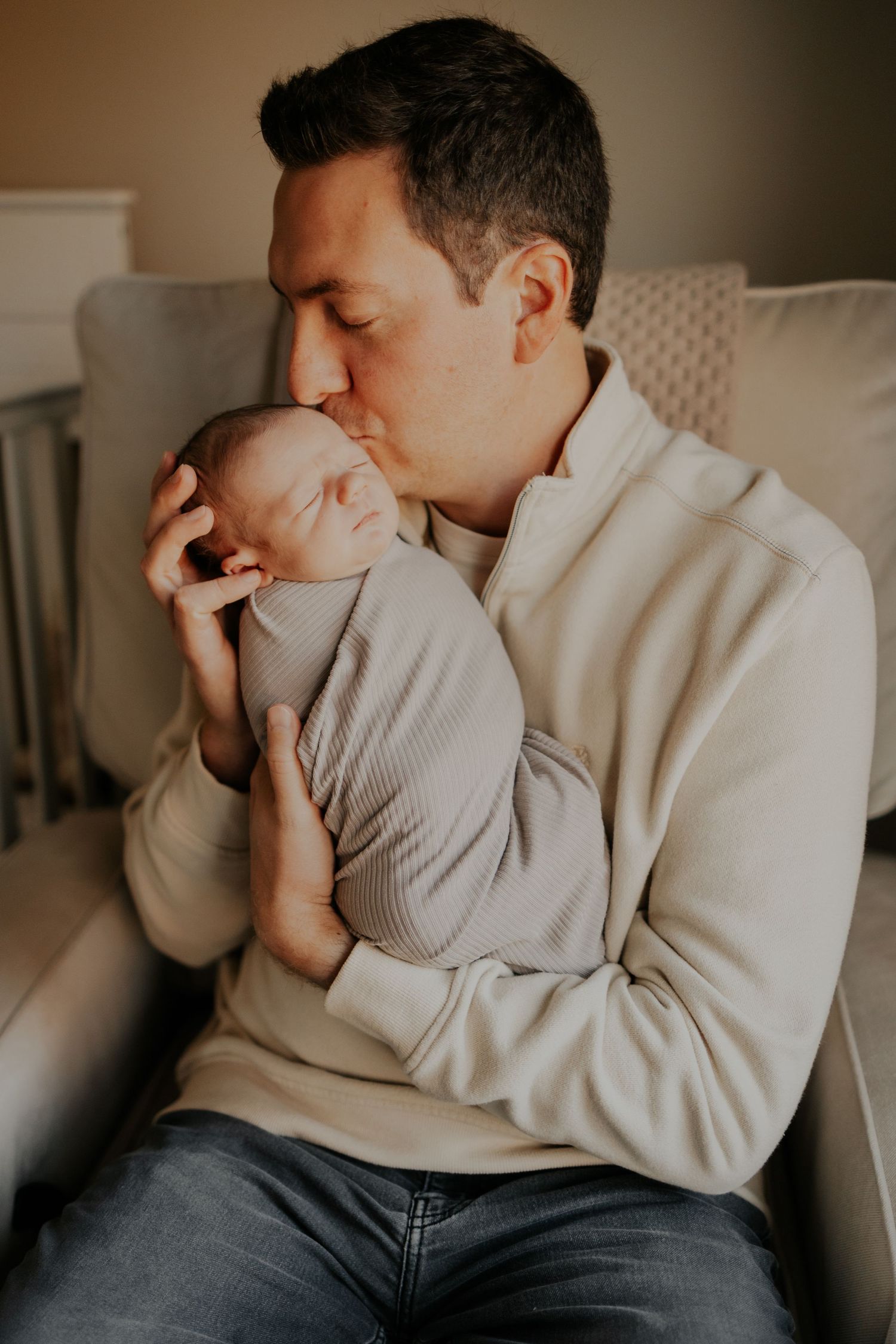 A tender moment showing a parent in a beige sweater gently holding and kissing a sleeping newborn baby.