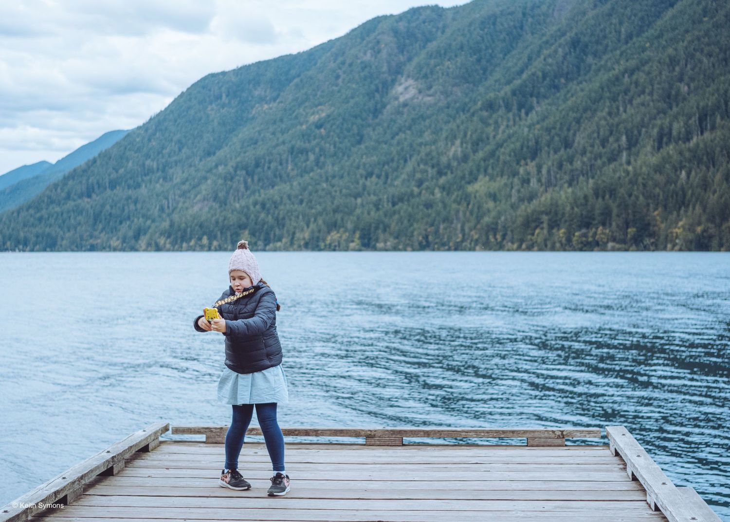 Someone takes photos from a wooden dock overlooking a scenic mountain lake with evergreen forests.
