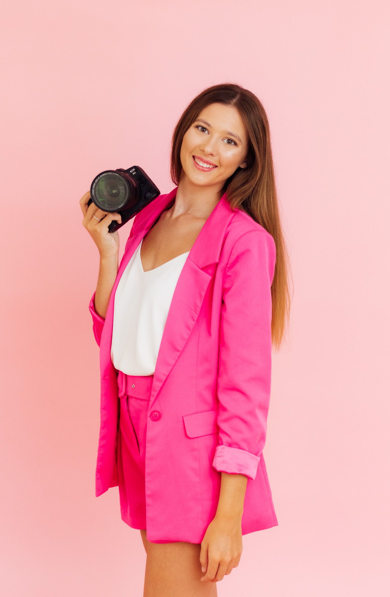 Professional photographer wearing vibrant pink blazer suit holding camera against pink backdrop.