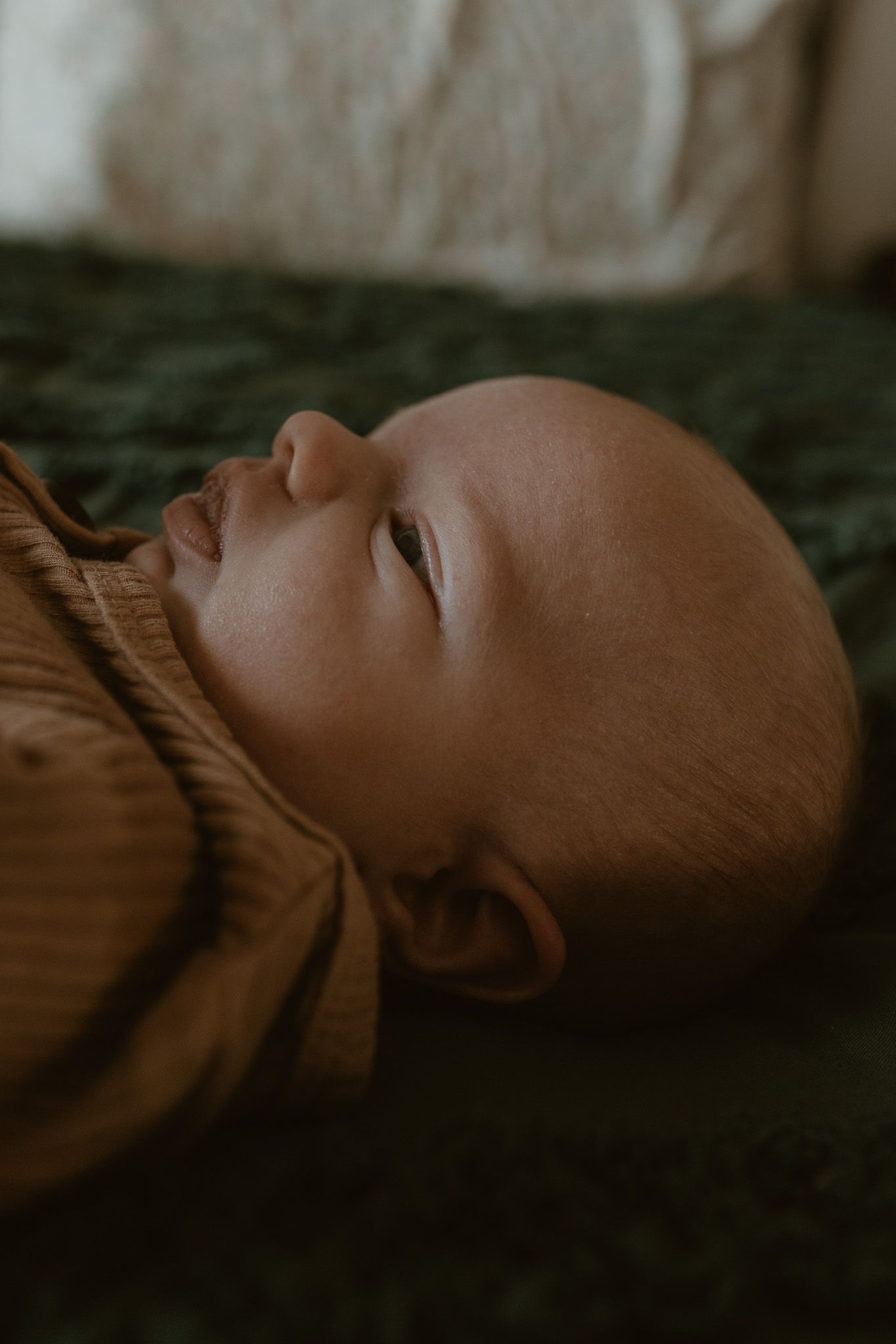 A sleeping baby in a brown shirt rests peacefully on a dark green textured surface in moody lighting.