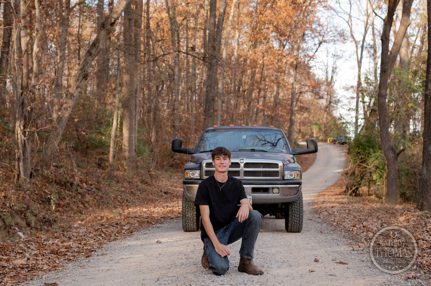 A person crouches in front of a black Dodge Ram pickup truck on a forest path.