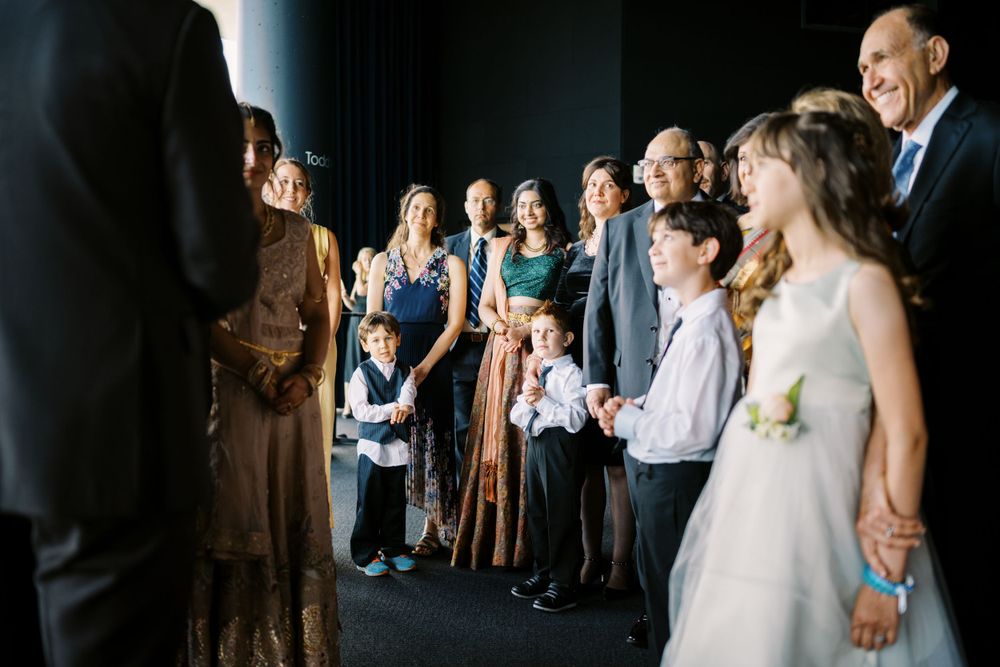 Wedding guests gather together and share joyful reactions as they watch something off-camera during a celebration.