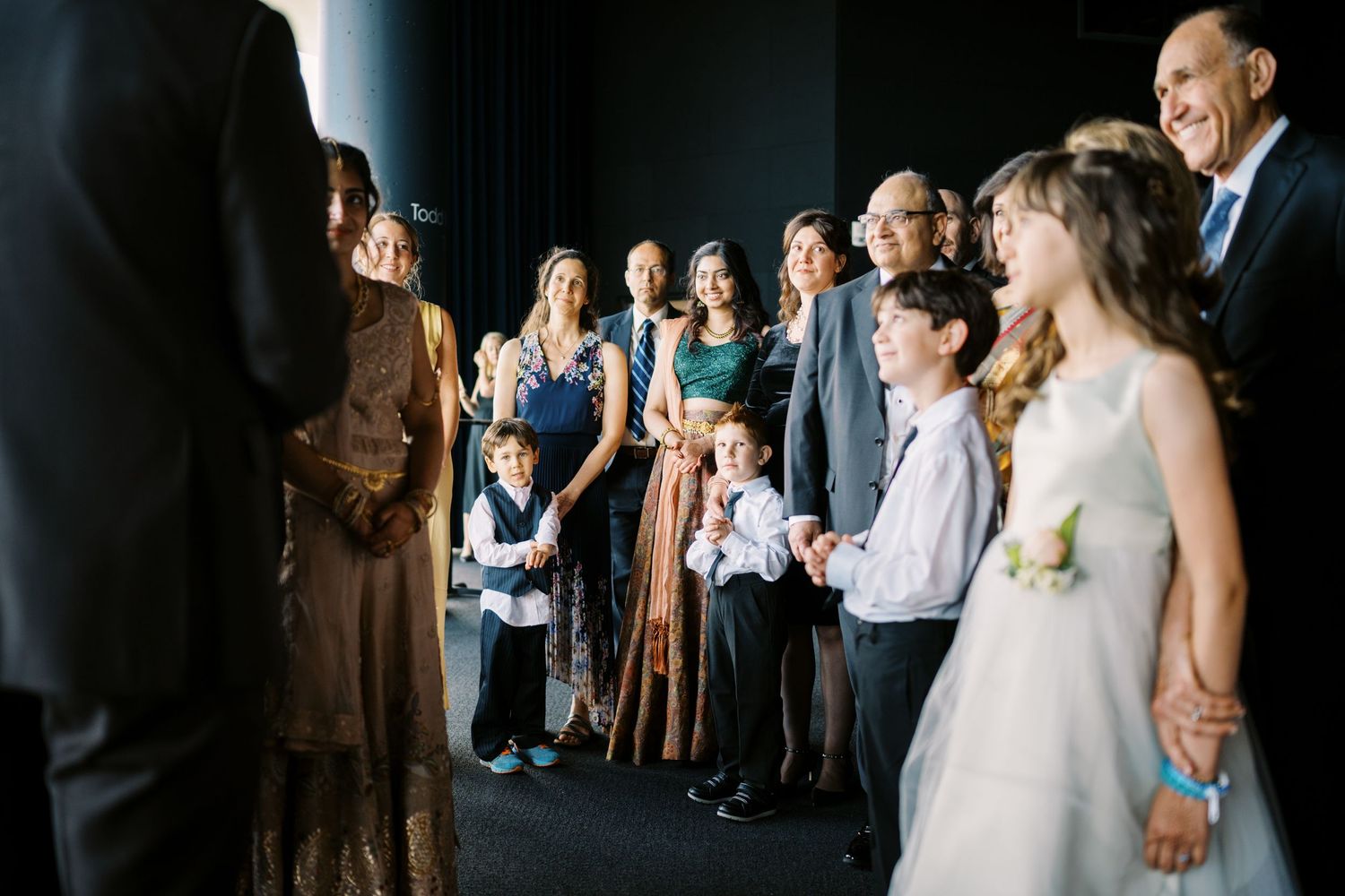 Wedding guests gather together and share joyful reactions as they watch something off-camera during a celebration.