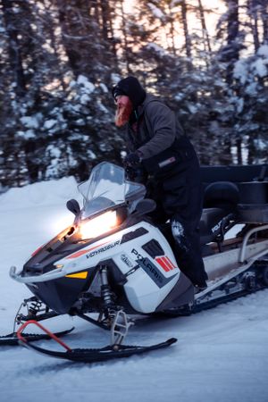 Snowmobile rider on white Polaris machine racing through snowy forest.