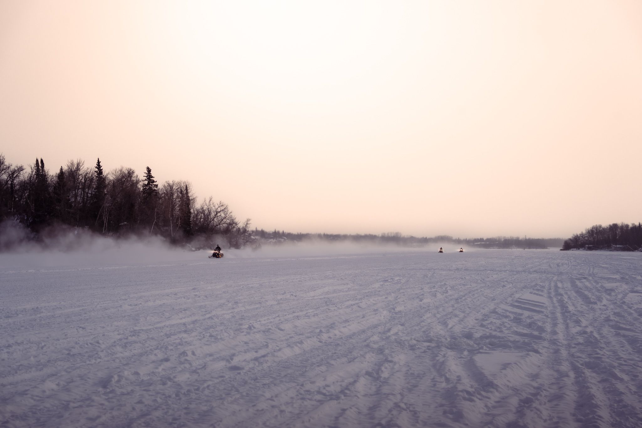 A snowy winter landscape with tracks across a frozen lake surrounded by evergreen forest in misty conditions.