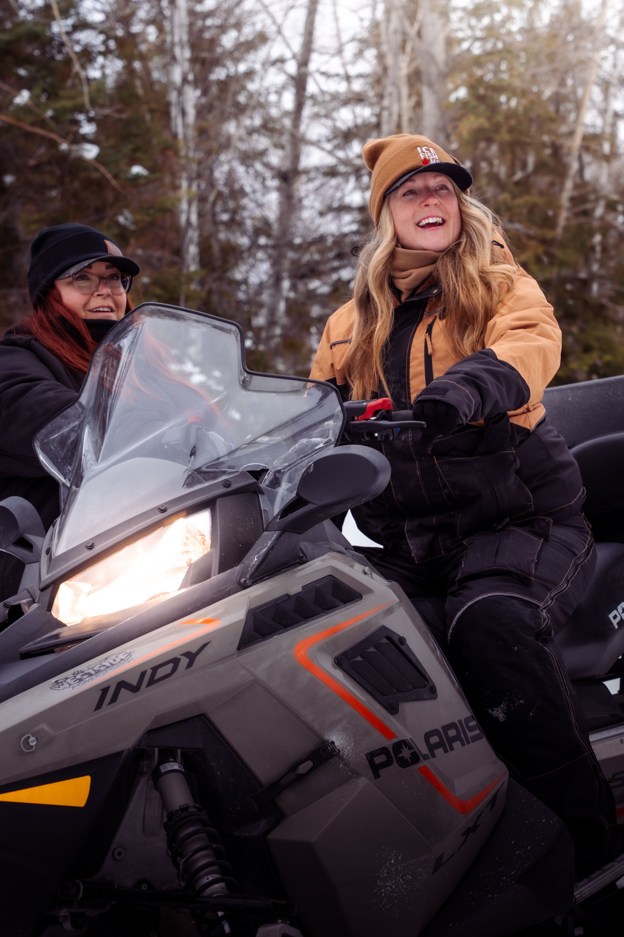 Two people enjoying a winter adventure on a Polaris Indy snowmobile in a snowy forest setting with headlights on.