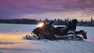 Black snowmobile parked on snowy terrain at sunset with forest scenery in the background.