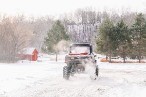 Off-road vehicle driving through snowy winter landscape with red barn in background.
