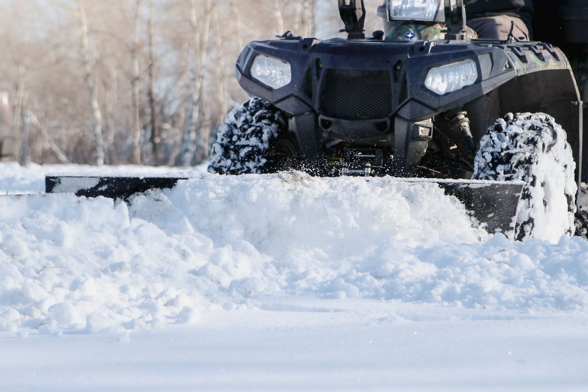Black ATV with snow plow attachment clearing fresh snow with bright LED headlights.