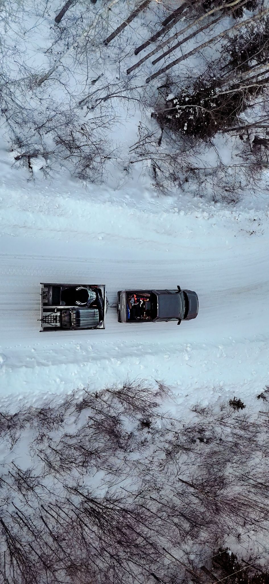 Aerial winter view of two parked vehicles on a snowy forest road surrounded by bare trees and white landscape.