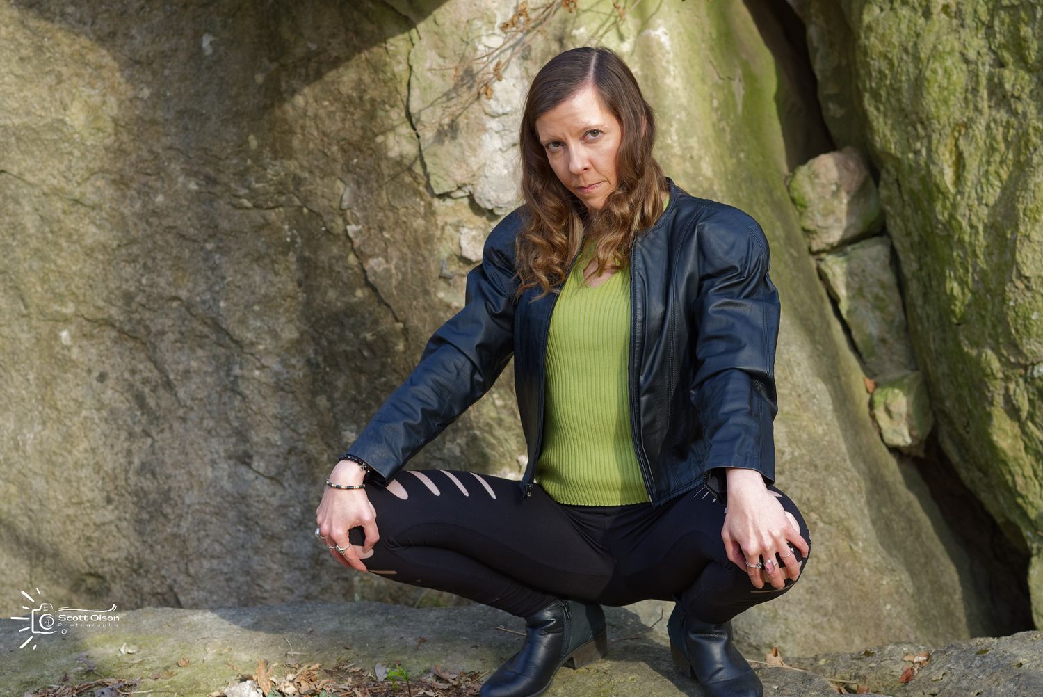 Model in black leather jacket and green sweater crouches on rocky outcrop against mossy stone wall.