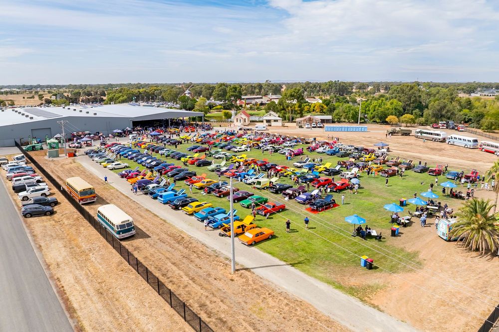 Aerial panorama of a large colorful car show displaying hundreds of vintage and custom vehicles on a grass field.