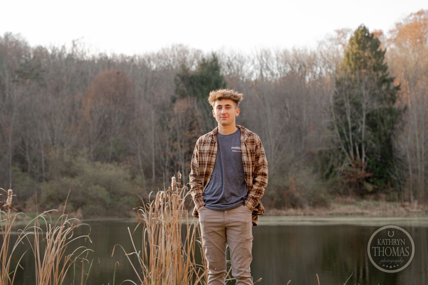 Someone in a plaid shirt stands by a serene lake with autumn trees in the background.