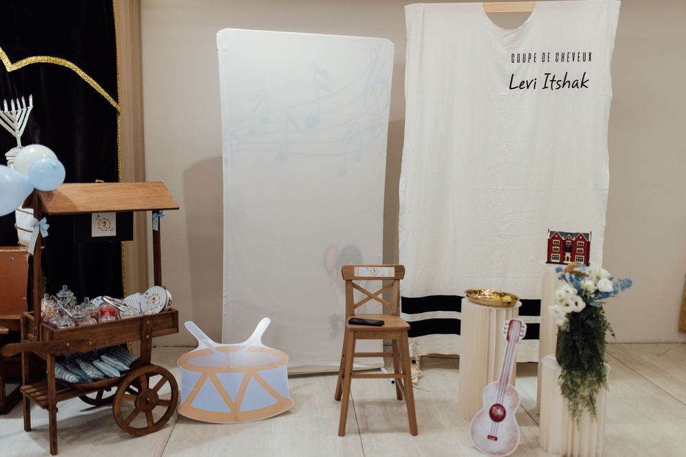 Jewish brit milah ceremony setup with white backdrop, wooden chair, and decorative cart with blue balloons.