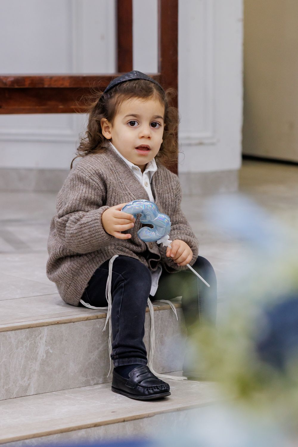 A small child in a brown sweater sits on wooden stairs while holding a blue stuffed toy.