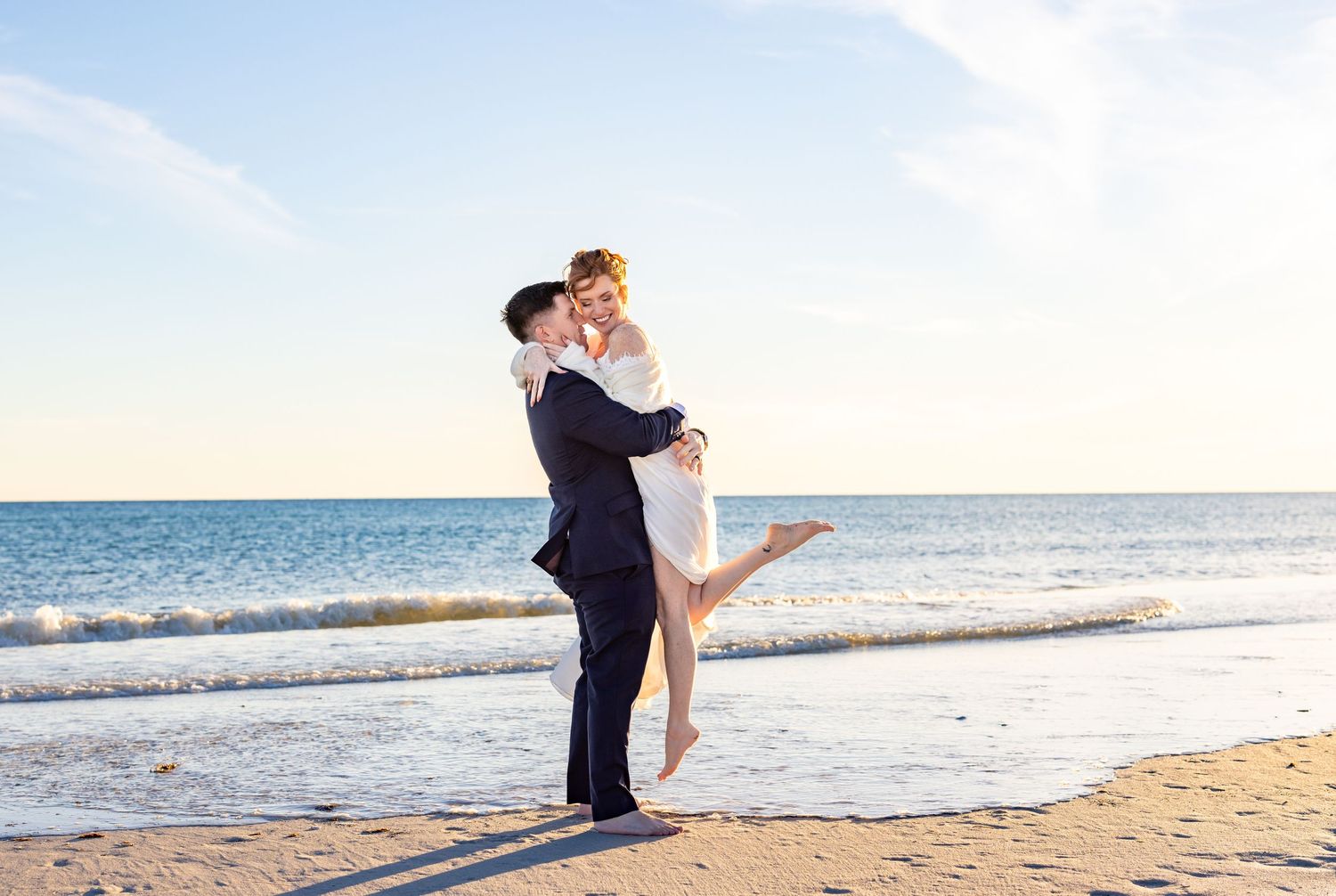 A couple embraces on a sunny beach with the ocean waves and horizon behind them during a romantic moment.