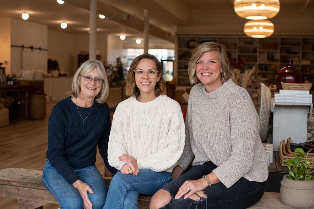 A brand photo of a small business team of three women smiling while sitting on a couch in coordinating cream sweaters and casual attire.