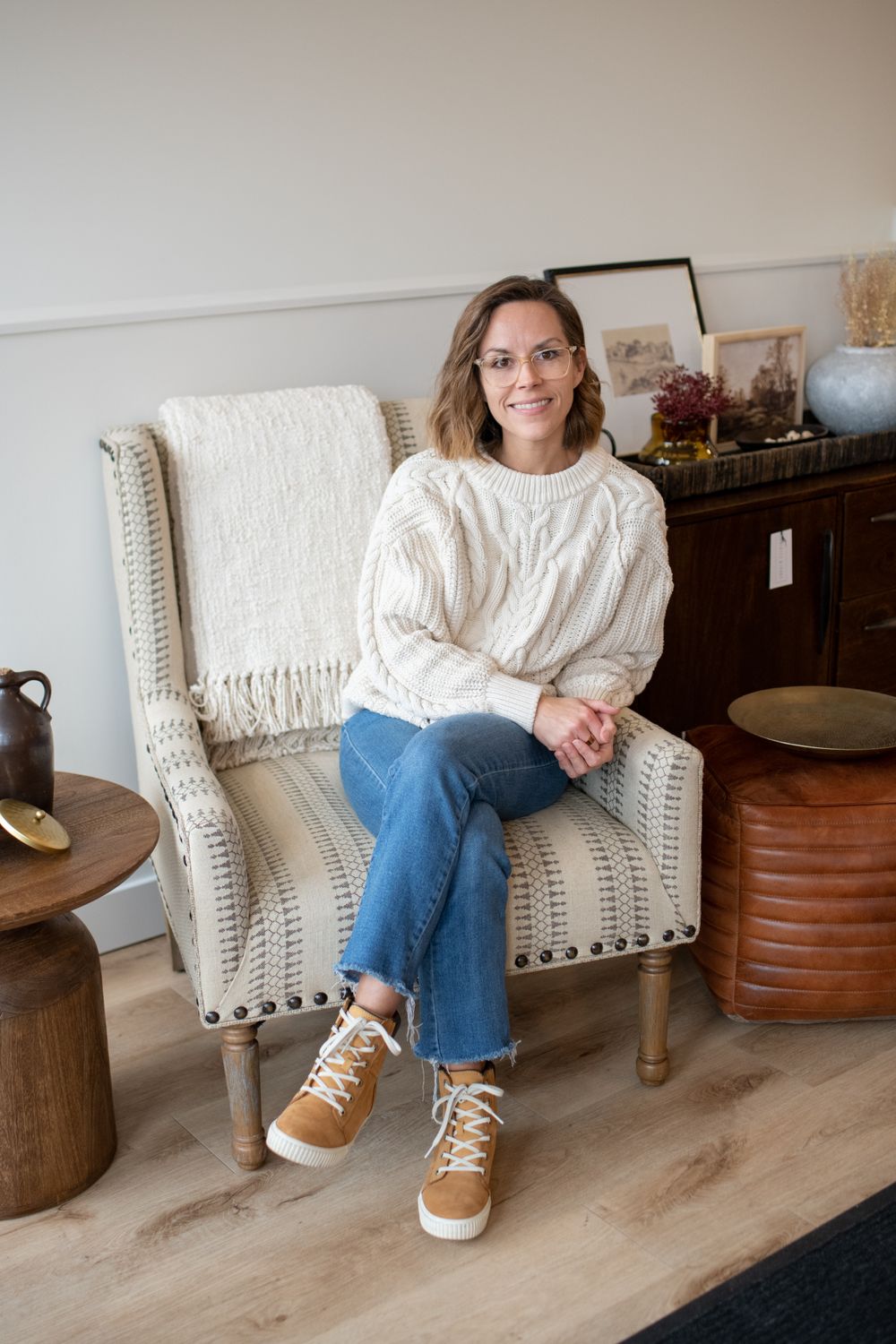 Person in white sweater and jeans sitting on upholstered chair with studded trim details.