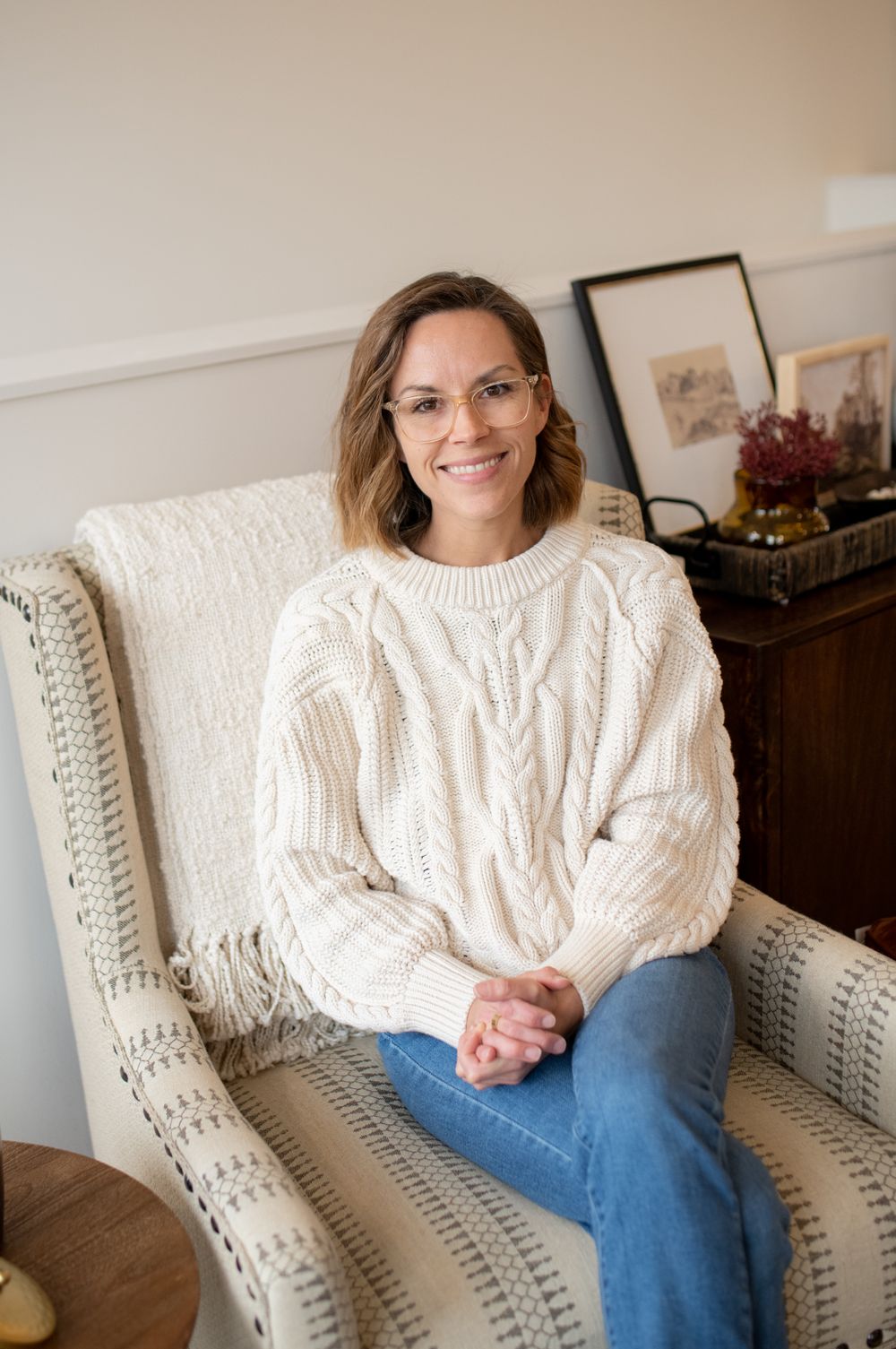 A brand photo of a woman business owner relaxing in cream cable knit sweater on light-colored accent chair.