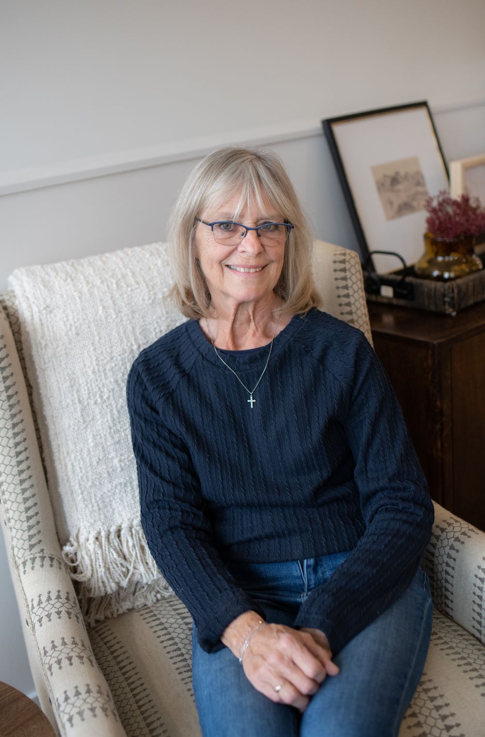 A brand photo headshot of a small business owner wearing navy sweater seated comfortably in a white upholstered armchair.