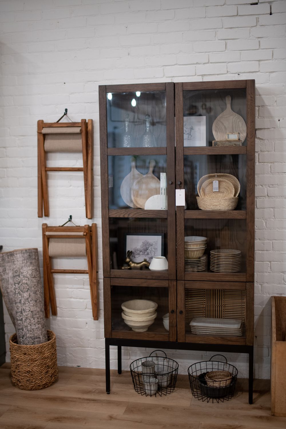 Glass display cabinets and wooden ladders arranged against white brick wall with woven baskets.