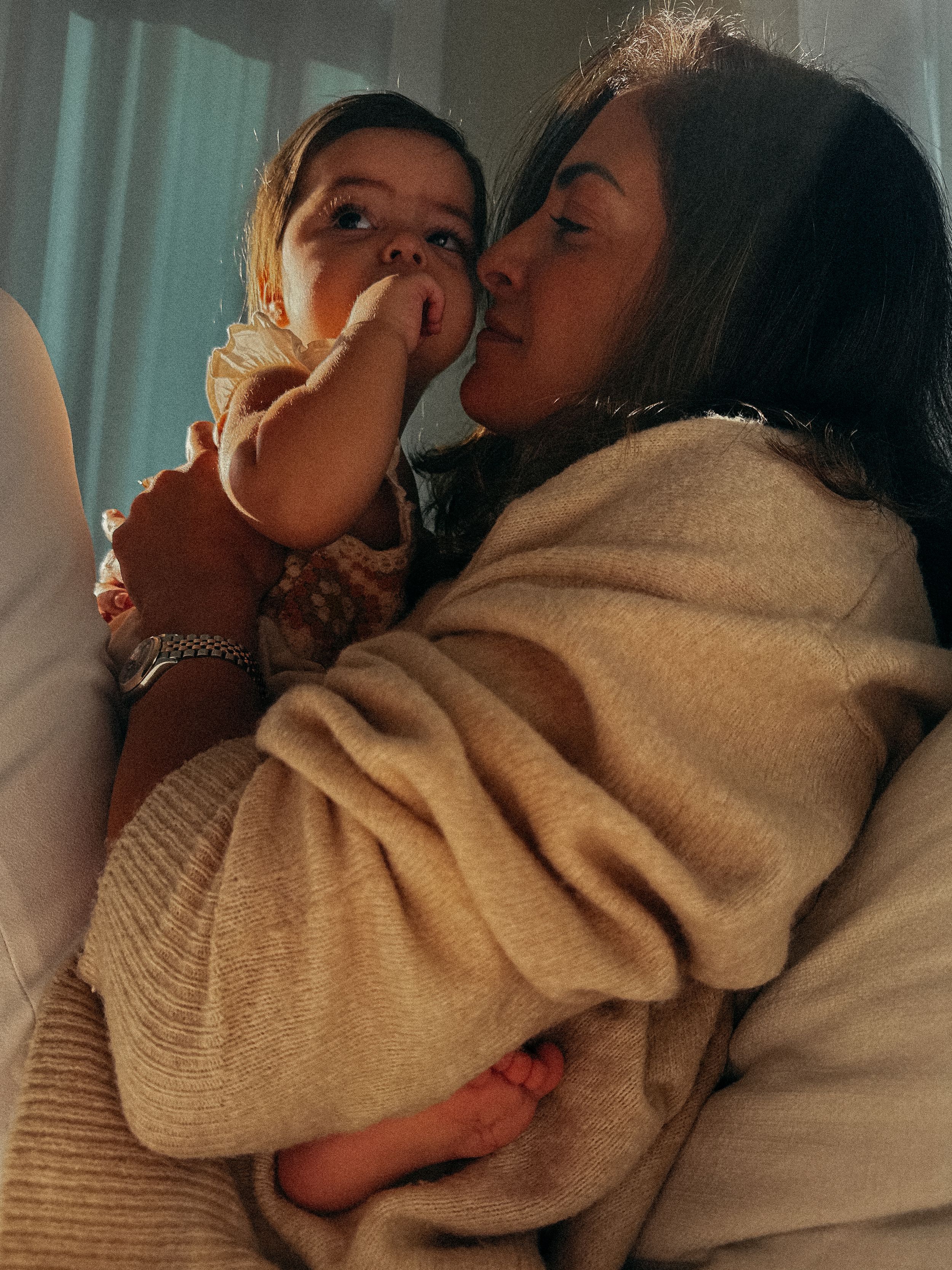 A woman dappled in sunlight lifts her baby up to her face and puts her nose on her babys cheek