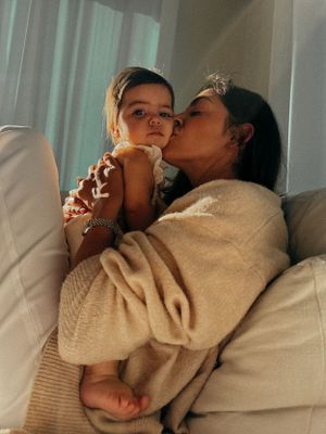 A woman dappled in sunlight lifts her baby up to her face and gives her a kiss on the cheek