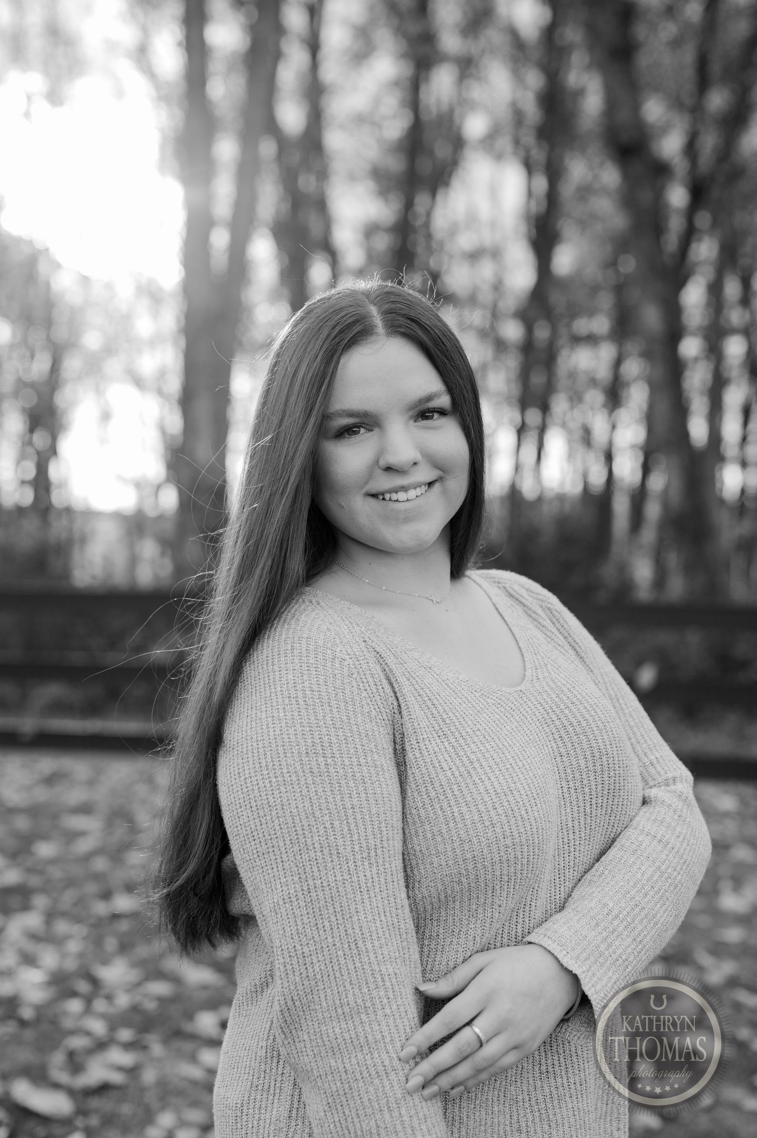 Black and white portrait of someone in a knit sweater against a wooded background with fallen leaves.