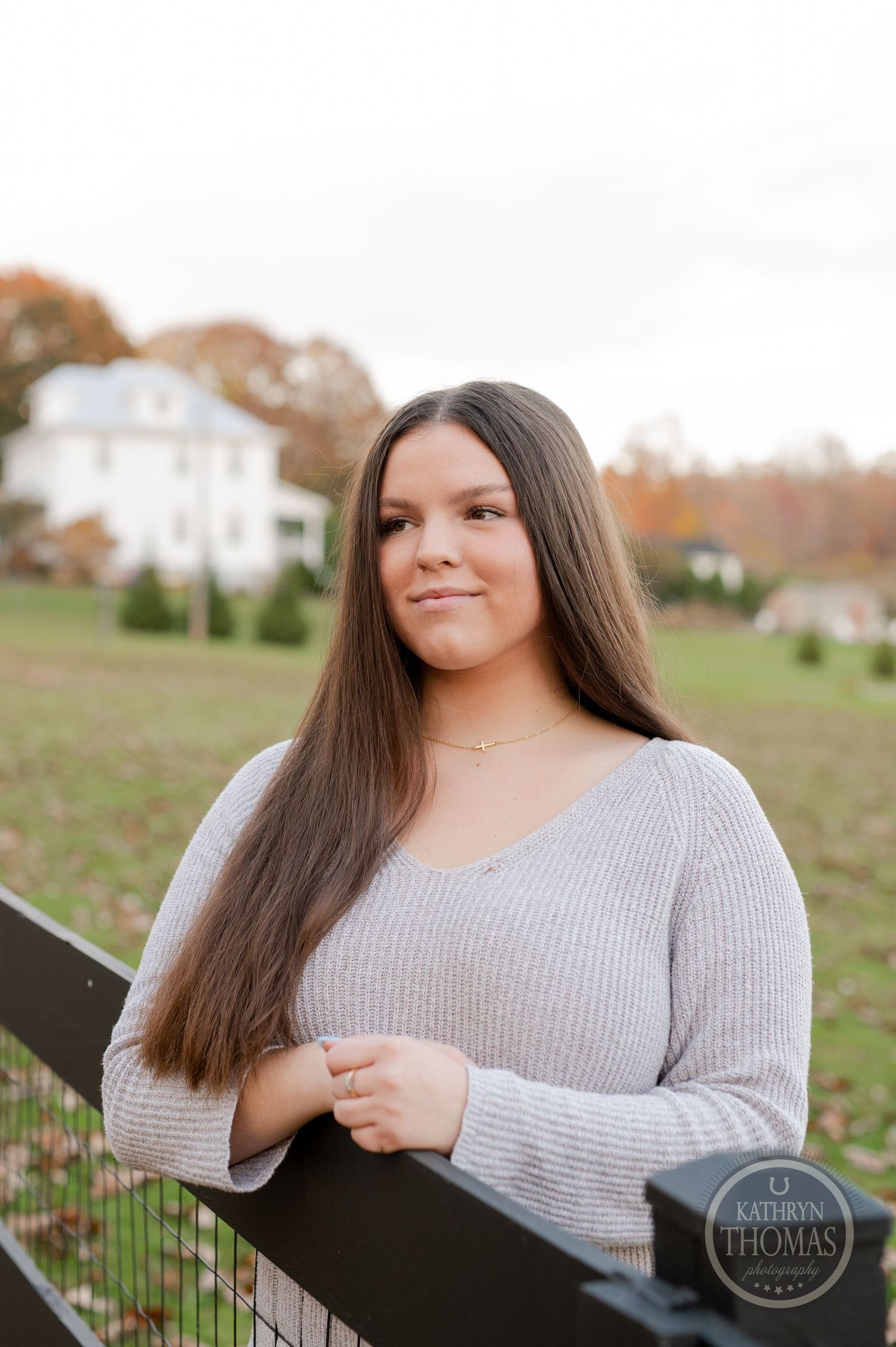 Natural autumn portrait by wooden fence with long straight hair and farmhouse backdrop.