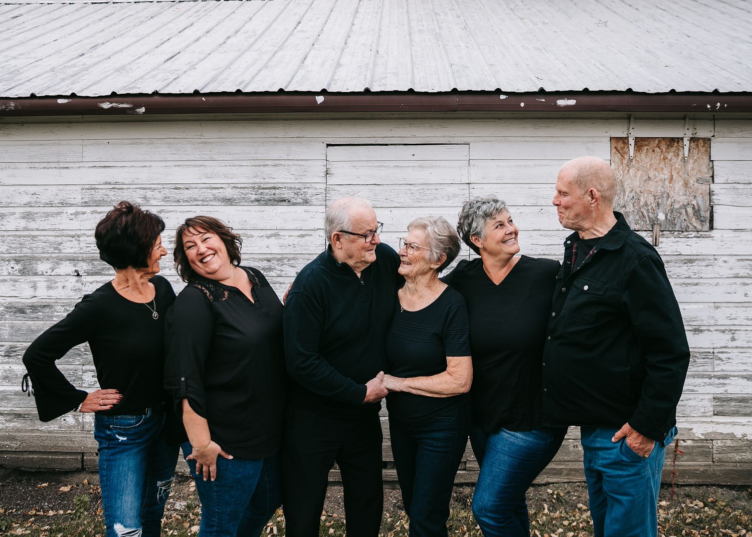 Group of six older adults in matching black outfits laughing together outside a rustic white barn.