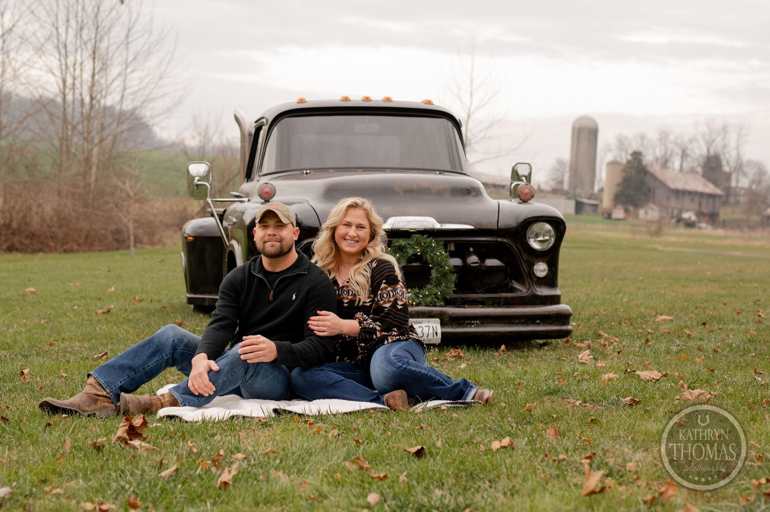 Couple relaxing on blanket in front of classic black truck in countryside setting.