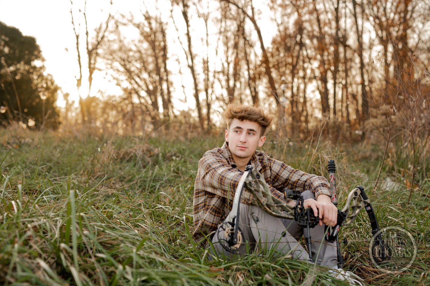 Archer relaxes in tall grass with compound bow during golden hour photo session.