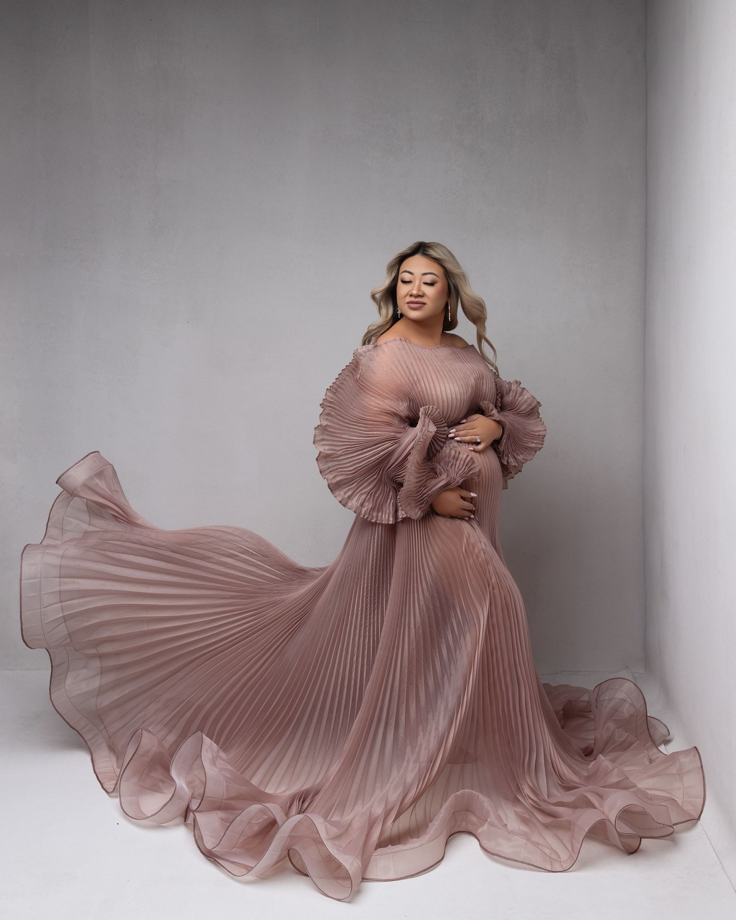 Ethereal maternity portrait with swirling dusty pink pleated dress creating elegant movement against neutral background.