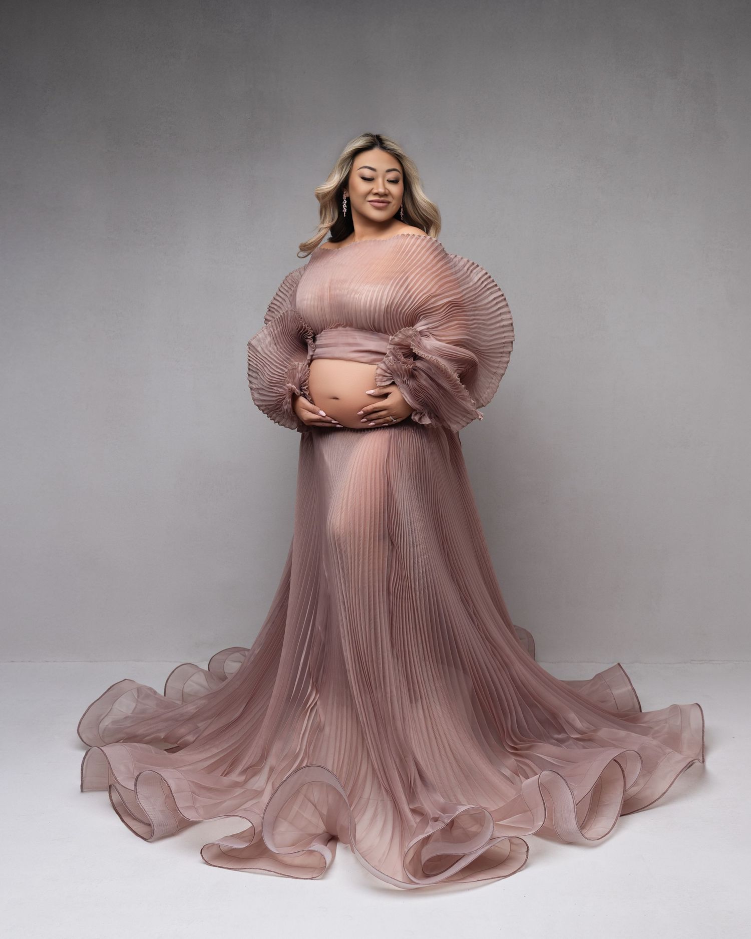 Maternity photoshoot featuring flowing dusty rose pleated gown with dramatic puff sleeves against gray backdrop.