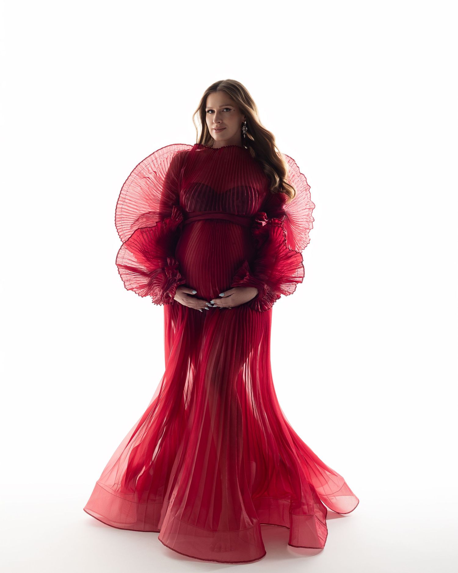 A figure in a flowing red tulle gown stands against a white backdrop while holding their abdomen in a maternity pose.