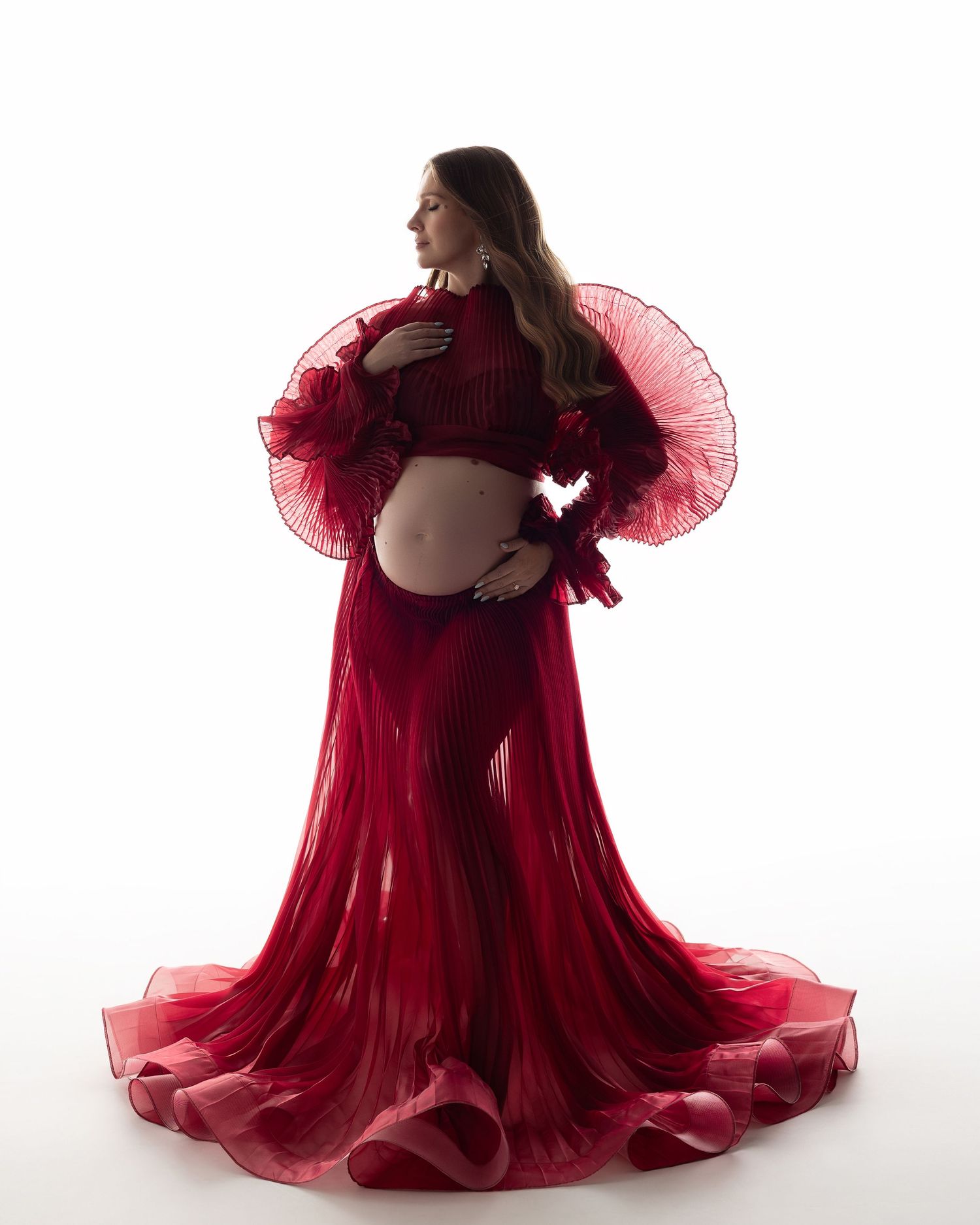A pregnant figure in a flowing red tulle gown poses gracefully against a white background in a maternity photoshoot.
