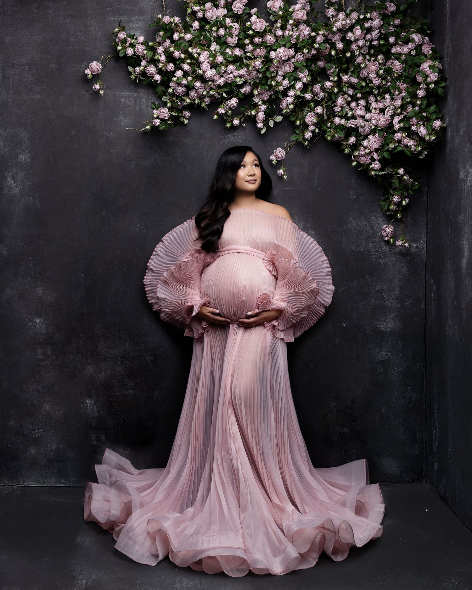 A pregnant figure in a flowing pink tulle gown poses against a dark wall adorned with delicate cherry blossoms.