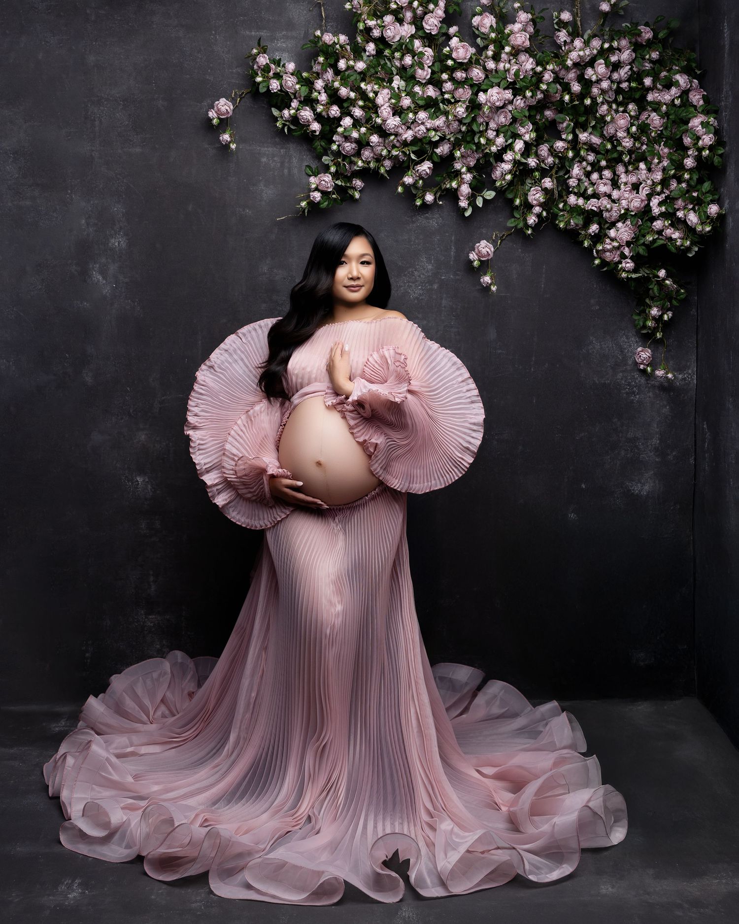 Elegant maternity photo with pink pleated gown and floral backdrop against dark grey wall.