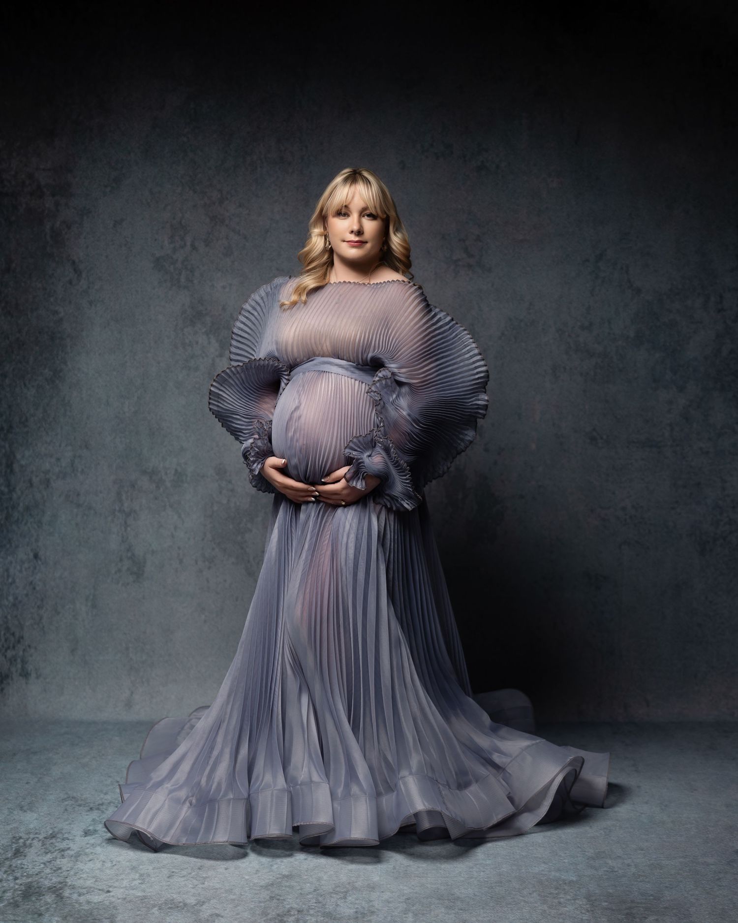 A pregnant figure in a flowing gray pleated gown poses against a dark textured backdrop in an elegant maternity portrait.