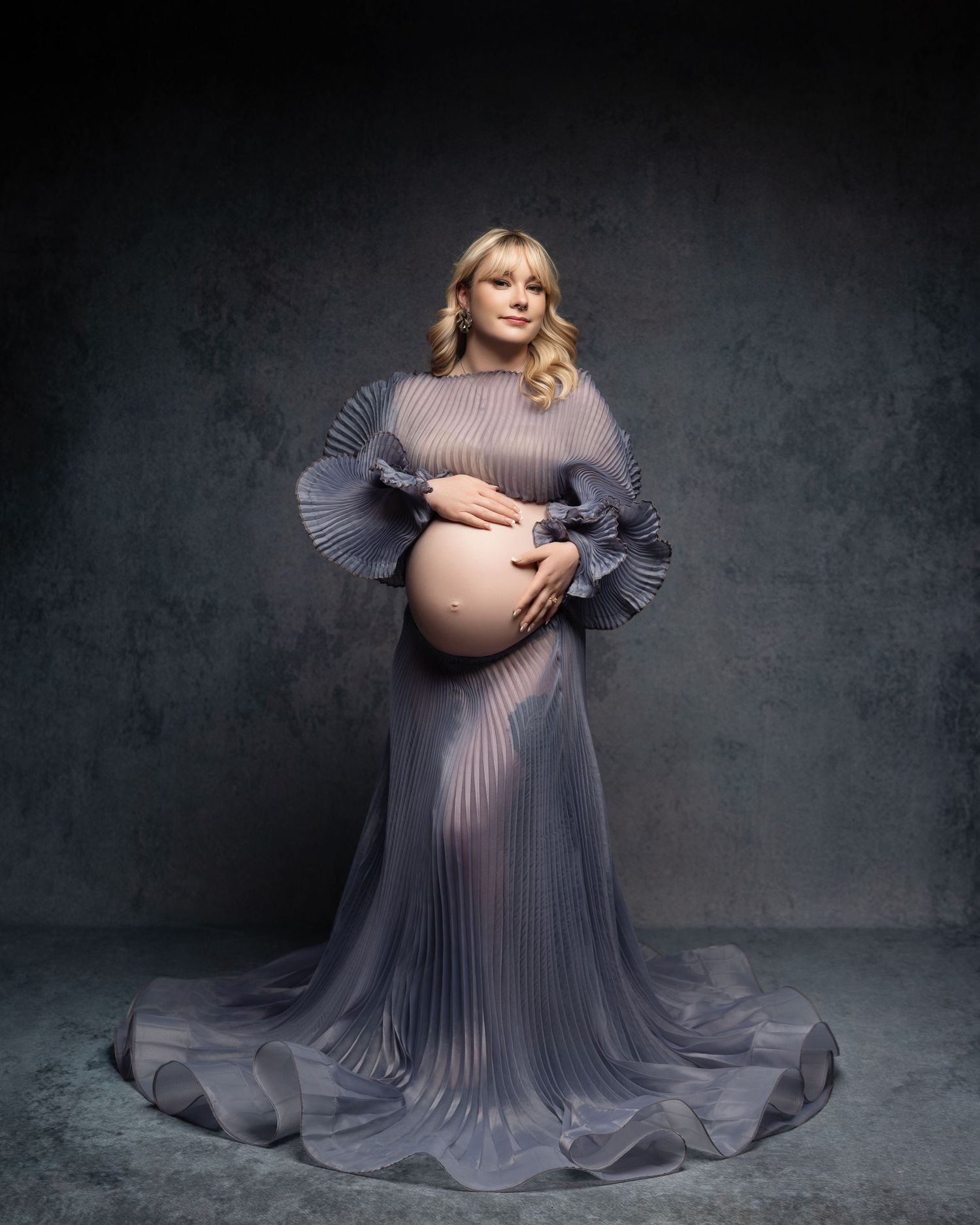 Ethereal maternity portrait features an elegant pleated gray gown with dramatic ruffled sleeves against a dark backdrop.