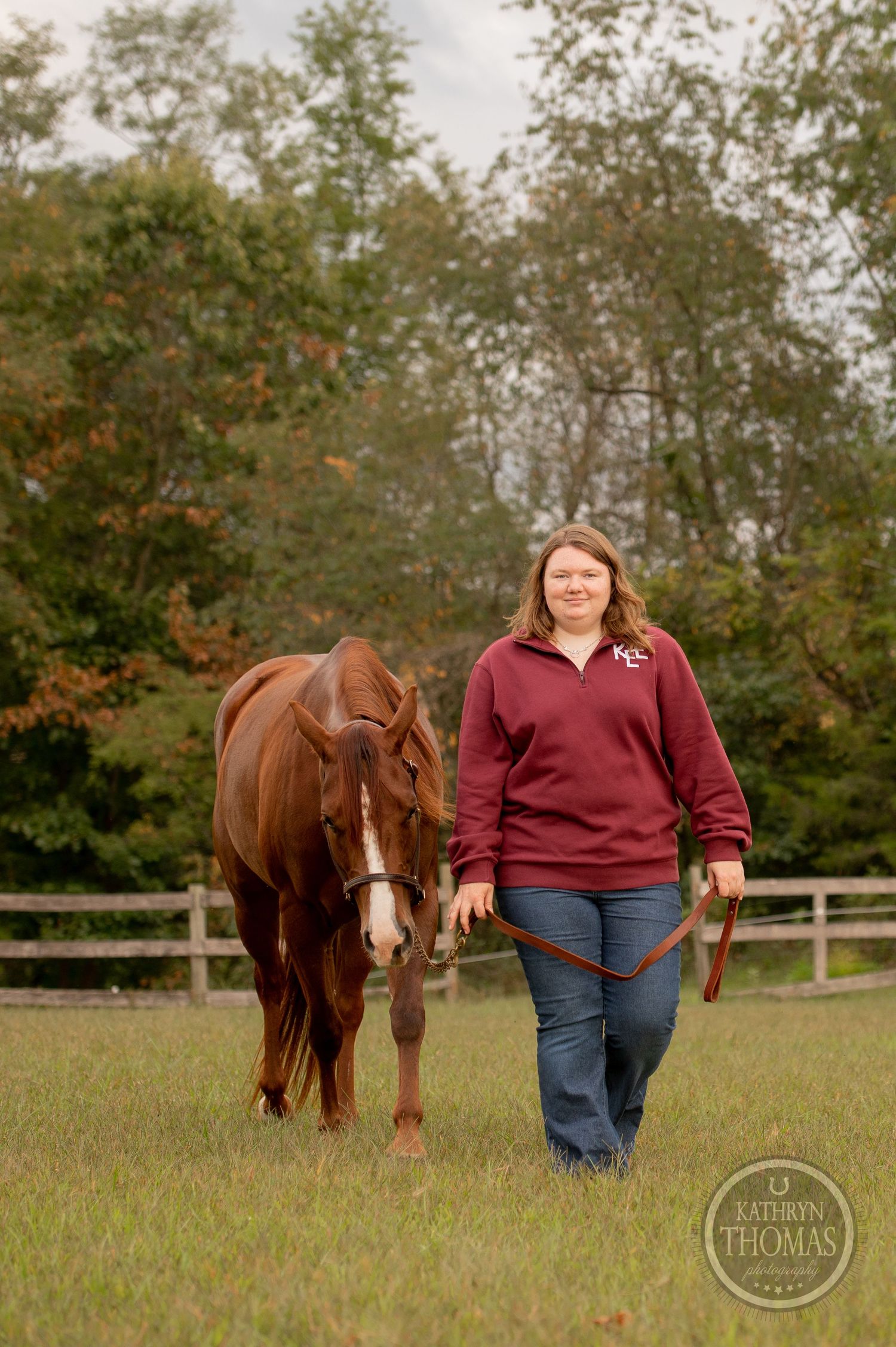 A chestnut horse follows alongside someone in a burgundy sweater through an autumn field.