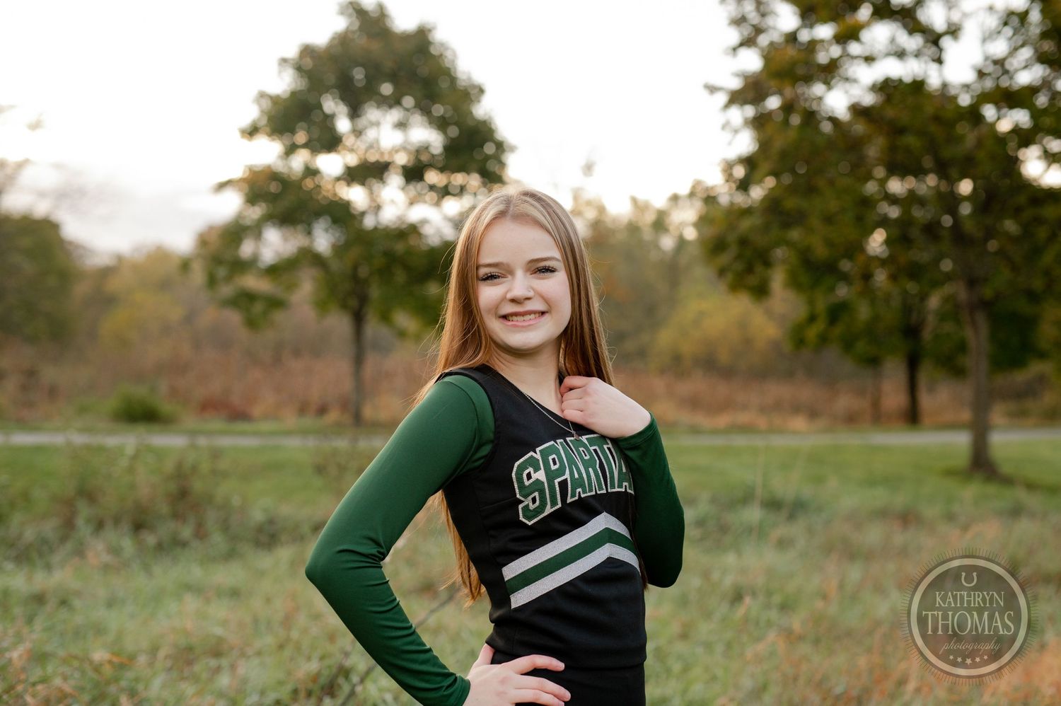 Cheerleader in green and black Spartans uniform poses against autumn field backdrop.