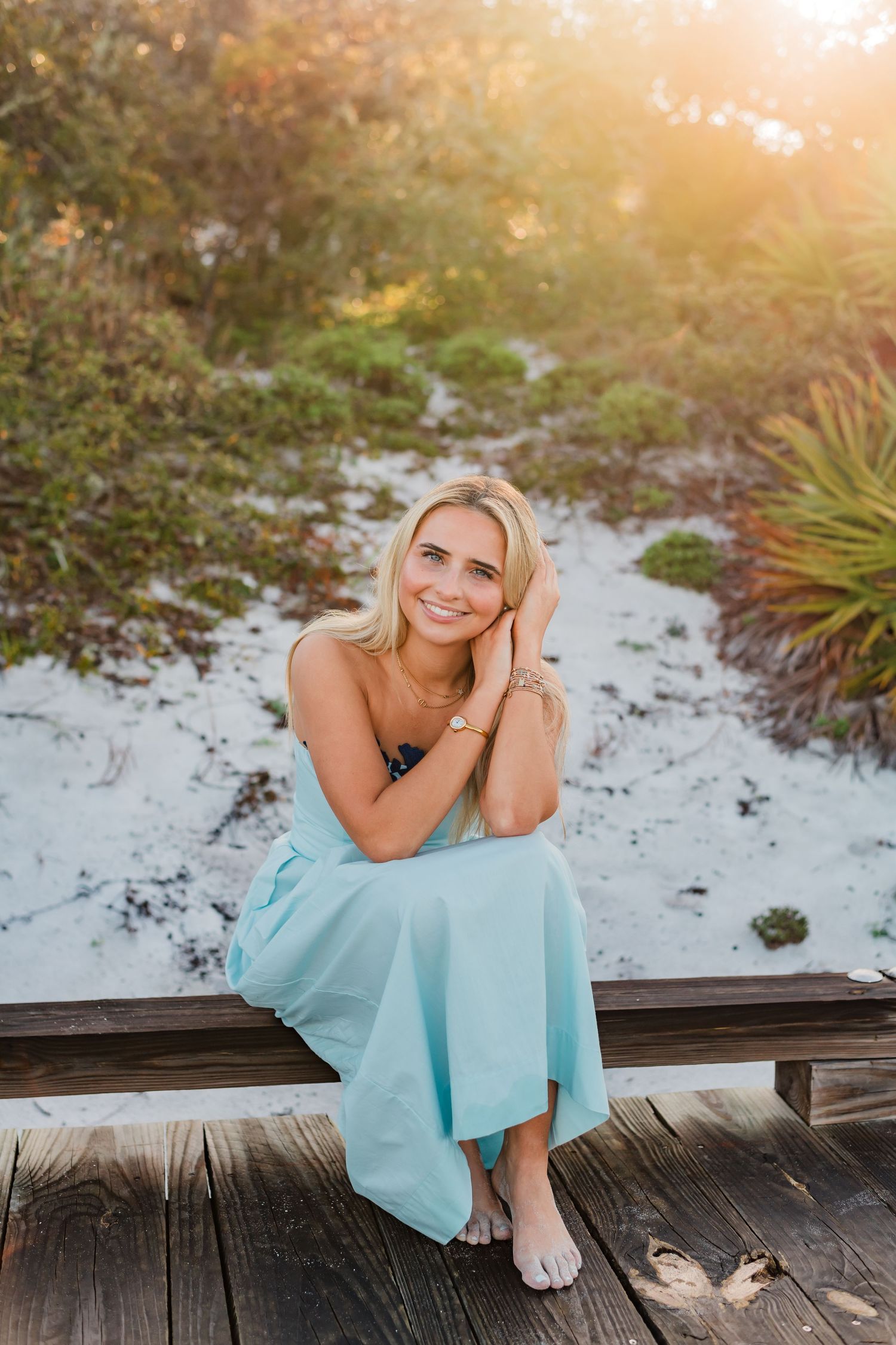 Smiling blonde woman in light blue dress sitting on wooden boardwalk near sandy beach with lush greenery backdrop.