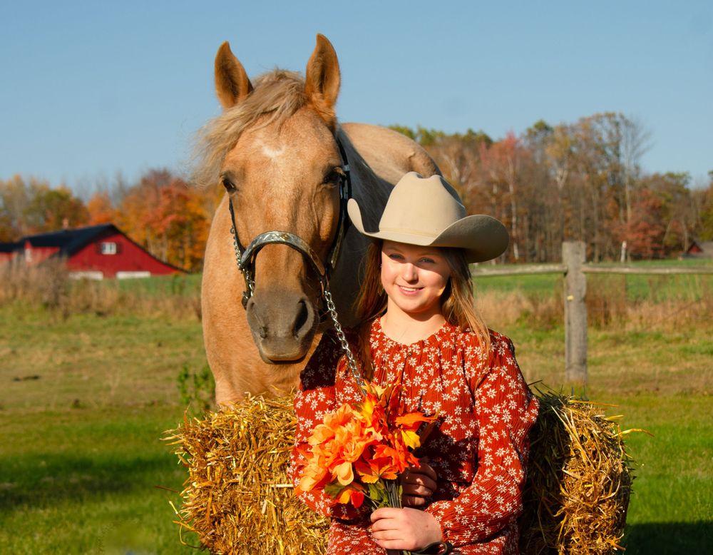Girl smiling at camera sitting on haybale with horse behind her looking at camera ears forward, in a Wisconsin fall photoshoot